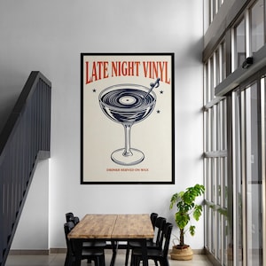 May include: A framed poster with the text "LATE NIGHT VINYL" in red and a graphic of a cocktail glass with a record inside. The poster also includes the text "DRINKS SERVED ON WAX" and is displayed in a room with a table and chairs.