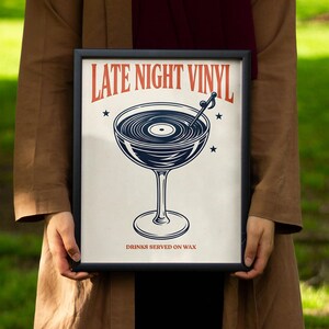 May include: A framed print with a black border featuring a cocktail glass design. The glass is filled with a vinyl record, and the text "LATE NIGHT VINYL" is displayed above. The phrase "DRINKS SERVED ON WAX" is below the glass.