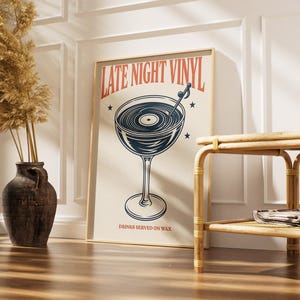 May include: A framed poster with the text "LATE NIGHT VINYL" in red and a graphic of a cocktail glass with a vinyl record inside. The poster also includes the text "DRINKS SERVED ON WAX" in a serif font. The poster is leaning against a white wall.