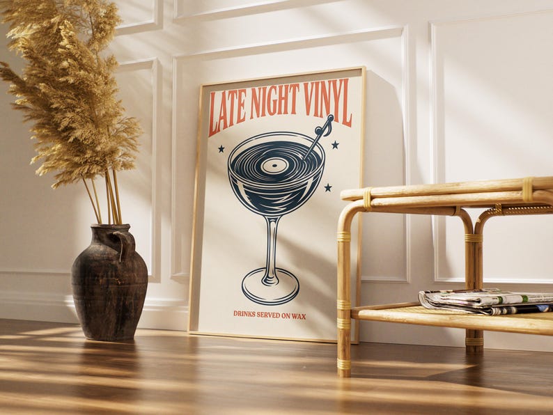 May include: A framed poster with the text "LATE NIGHT VINYL" in red and a graphic of a cocktail glass with a vinyl record inside. The poster also includes the text "DRINKS SERVED ON WAX" in a serif font. The poster is leaning against a white wall.