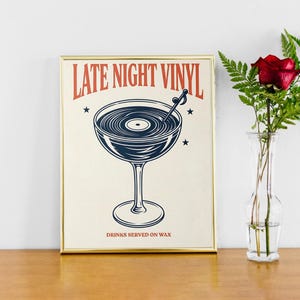 May include: Framed print with a vintage-style illustration. The artwork features a cocktail glass filled with a vinyl record design, with the text "LATE NIGHT VINYL" in red and "DRINKS SERVED ON WAX" below. A red rose in a glass vase is next to the framed print.