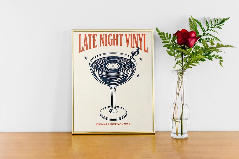 May include: Framed print with a vintage-style illustration. The artwork features a cocktail glass filled with a vinyl record design, with the text "LATE NIGHT VINYL" in red and "DRINKS SERVED ON WAX" below. A red rose in a glass vase is next to the framed print.