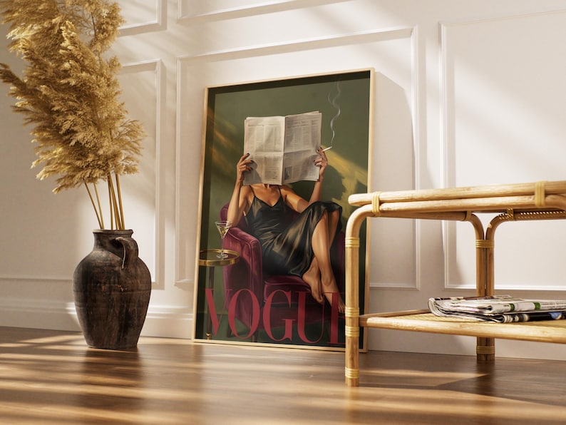 May include: A framed art print featuring a woman in a black dress, seated in a red velvet chair, reading a newspaper with the word "VOGUE" in large red letters. A martini glass sits on a small table. A decorative vase with dried plants is on the left.