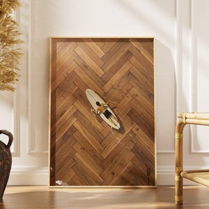 May include: A rectangular wall art print featuring a top-down view of a person lying on a white surfboard, set against a background of brown wooden planks arranged in a herringbone pattern. The print is framed in a thin, light-colored wood frame.