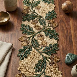 May include: Beaded table runner featuring a nature-inspired design. The runner showcases a cream-colored background with a pattern of green leaves and golden acorns. The runner measures approximately 14 inches wide and 72 inches long.