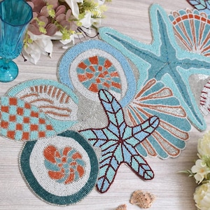 Puede incluir: Decoraciones de mesa con cuentas de temática costera, con diseños de estrellas de mar y conchas marinas. Los artículos utilizan cuentas azules, naranjas y blancas en patrones intrincados. También hay un vaso azul y flores.