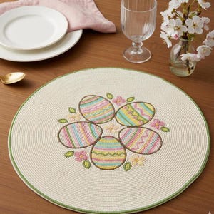 Può includere: Un sottopentola rotondo color crema con un bordo verde. Il centro presenta un disegno ricamato di cinque uova di Pasqua colorate circondate da piccoli fiori e foglie verdi. Il sottopentola è appoggiato su un tavolo di legno, con un piatto, un bicchiere e dei fiori sullo sfondo.