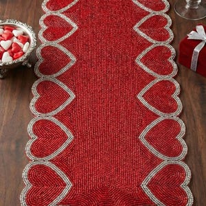 Może przedstawiać: Czerwony, koralikowy bieżnik na stół z motywami w kształcie serca obrysowanymi srebrem. Bieżnik jest ozdobiony czerwoną różą, srebrnym świecznikiem i małą miseczką z cukierkami w kształcie serca. Obecne jest również czerwone pudełko na prezent.