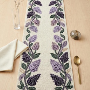 May include: Cream-coloured beaded table runner with a floral design. The runner features two vertical rows of purple and green beaded flowers and leaves. A gold spoon, napkin, and decorative stone are also visible.