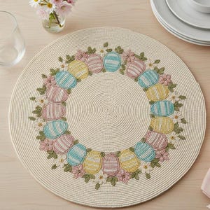 Könnte beinhalten: Runder, cremefarbener Platzdeckchen mit einem Osterei- und Blumendesign. Die Eier sind in Pastellfarben gehalten, und die Blumen sind rosa und weiß. Der Platzdeckchen besteht aus gewebtem Material.