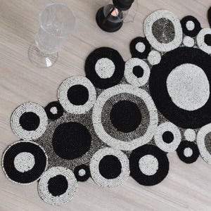 May include: A beaded table runner with a circular design in black, white, and gray. The circles vary in size and are arranged in a flowing pattern. A clear glass and a black lantern are also visible.
