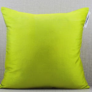 May include: A square, lime green pillow with a soft, textured appearance. The pillow is centered in the frame, with a tag attached to the upper right corner. The background is a light gray, textured surface.