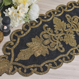 Może przedstawiać: Czarny, koralikowy bieżnik na stół ze złotymi wzorami kwiatowymi i wiciami. Bieżnik ma falisty brzeg i jest ozdobiony złotymi koralikami. W tle znajdują się przedmioty dekoracyjne, w tym złoty wazon i białe kwiaty.