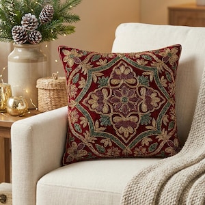 Pode incluir: Uma almofada quadrada decorativa com um design floral bordado em ouro, verde e bordô sobre um fundo vermelho. A almofada está colocada numa poltrona creme, com uma manta de tom neutro sobre o braço. O design é ornamentado.