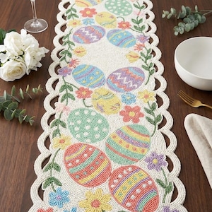 May include: Beaded Easter table runner featuring colorful Easter eggs and floral designs. The runner has a scalloped edge and is decorated with pastel colors, including pink, yellow, blue, and green. The runner is approximately 60 inches long.