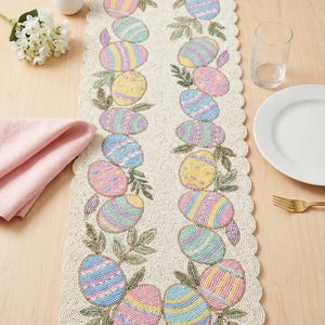May include: Beaded Easter table runner with scalloped edges. The runner features pastel-coloured Easter eggs with striped and dotted patterns, surrounded by green leaves. The runner is on a light-coloured wooden table.