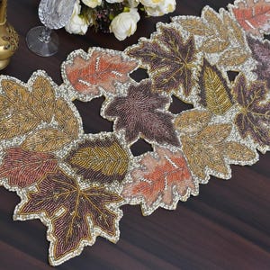 Può includere: Runner da tavolo con perline con un design a foglie nei toni dell'oro, del marrone e dell'arancione. Le foglie sono finemente dettagliate e delineate con un colore più chiaro, creando un aspetto strutturato. Ideale per l'arredamento autunnale o stagionale.