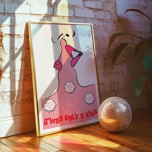 May include: A framed art print with a gold border, featuring a pink illustration of a mouth and the text "IT WAS ONLY A KISS". A disco ball sits on the wooden floor. The artwork leans against a white brick wall.