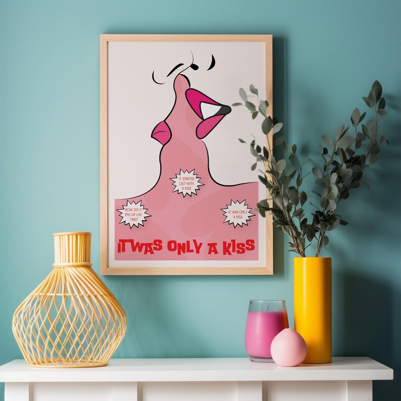 May include: A framed art print featuring a pink illustration of kissing lips. The print includes the text "IT WAS ONLY A KISS" and speech bubbles. A yellow vase and candle are on a white surface.