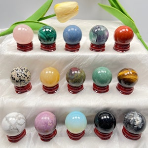 May include: A collection of 15 polished gemstone spheres displayed on small wooden stands. The spheres are various colours, including pink, green, blue, red, yellow, brown, black, white, and grey.