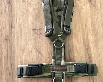 May include: A green camouflage dog harness with black buckles and a silver D-ring. The harness has a brown label that reads "Dander Wear" with a heart symbol.