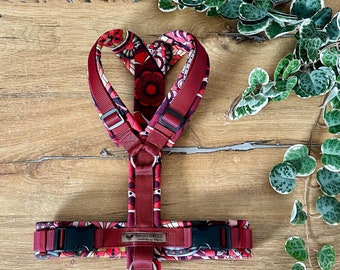 May include: A red dog harness with a floral pattern and black buckles. The harness has a metal ring for attaching a leash.