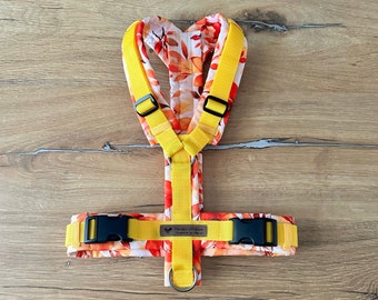 May include: Yellow dog harness with a white and orange floral pattern. The harness has a black buckle and a silver D-ring. The text "Pawsitive Vibes" is printed on the harness.