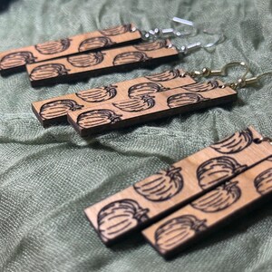May include: Wooden earrings with a rectangular shape and a repeating pattern of a fruit design. The earrings have silver-colored hooks. The earrings are displayed on a green fabric surface.