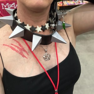 May include: A black leather choker with silver spikes and star accents. A silver pentagram pendant hangs from a red lanyard. The person is wearing a black vest top.