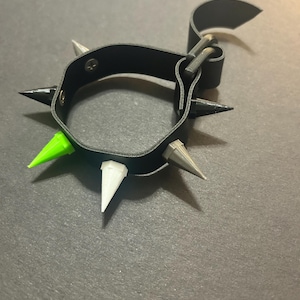 May include: Black leather wristband with multiple spikes in various colors, including green, white, and gray. The wristband has a buckle closure and a loop. The spikes are evenly spaced around the band.