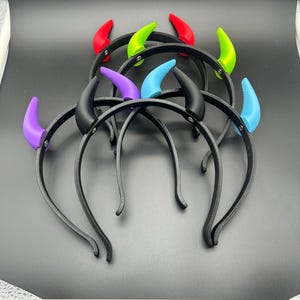 May include: A collection of black headbands, each featuring a pair of devil horns in various colors. The horns are red, green, blue, purple, and black. The headbands are arranged on a dark surface.