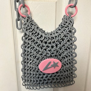 Puede incluir: Bolso gris estilo cota de malla con detalles en rosa. El bolso presenta un emblema ovalado rosa con una estrella gris y una letra estilizada. El bolso tiene una correa de cadena gris y asas circulares rosas.