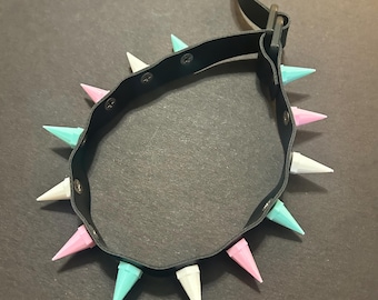 May include: A black leather choker with a silver buckle and a row of colorful spikes. The spikes are alternating colors of pink, white, and light blue.