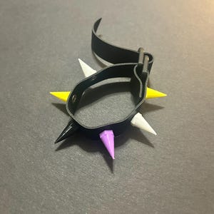 May include: A black leather bracelet with adjustable buckle closure and multiple spikes in white, yellow, and purple.