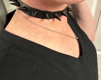 May include: A black leather choker with silver spikes. The choker is worn by a person with a silver nose ring.