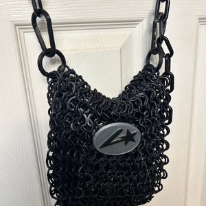 Peut inclure: Un sac en maille de chaîne noire avec une bandoulière en chaîne noire et un logo ovale gris avec une étoile noire et un éclair.