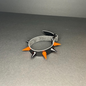Puede incluir: Una pulsera de cuero negro con púas naranjas y negras. La pulsera tiene un interior gris y una correa negra con cierre de hebilla. Las púas están espaciadas uniformemente alrededor de la pulsera.