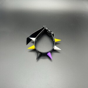 Puede incluir: Una pulsera negra que presenta una serie de púas puntiagudas en colores alternos amarillo, blanco, negro y morado. La banda está construida con un material liso e incluye un cierre de hebilla de plástico negro para asegurar la forma circular.