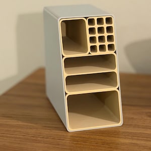 May include: A rectangular, multi-compartment desk organizer. The organizer has a light gray exterior with a beige interior. It features a large open compartment, two shelves, and a grid of small square compartments. The organizer is made of a sturdy material.