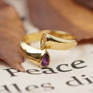 May include: A gold ring featuring a purple gemstone and a smaller clear gemstone. The ring is resting on a piece of paper with the text "Peace Pr...nd alles divis..." printed on it.