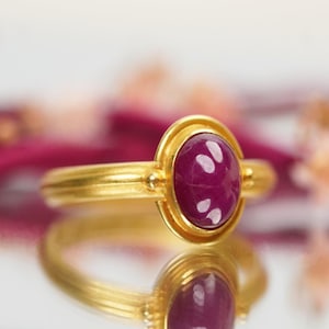 Puede incluir: Un anillo de oro con una piedra ovalada de color rojo intenso engastada en un engaste de bisel. El anillo tiene una banda texturizada y está acentuado con pequeñas cuentas de oro.