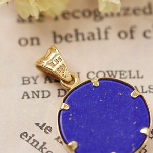 May include: A gold pendant with a round, deep blue stone. The pendant has a gold bail and a gold frame around the stone. The stone has a slightly textured surface. The background is a vintage paper with text.