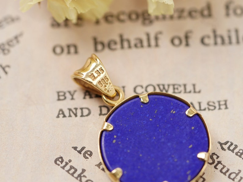 May include: A gold pendant with a round, deep blue stone. The pendant has a gold bail and a gold frame around the stone. The stone has a slightly textured surface. The background is a vintage paper with text.