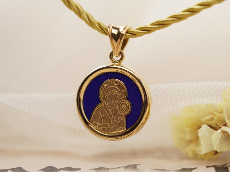 May include: Gold pendant necklace featuring a round pendant with a blue center and a gold religious icon. The pendant hangs from a gold-colored cord. The icon depicts a religious figure holding a child.