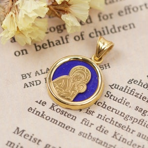 May include: Gold-tone pendant with a blue center featuring a religious icon. The pendant has a circular design with a detailed image of a religious figure. The pendant is on a vintage book page.