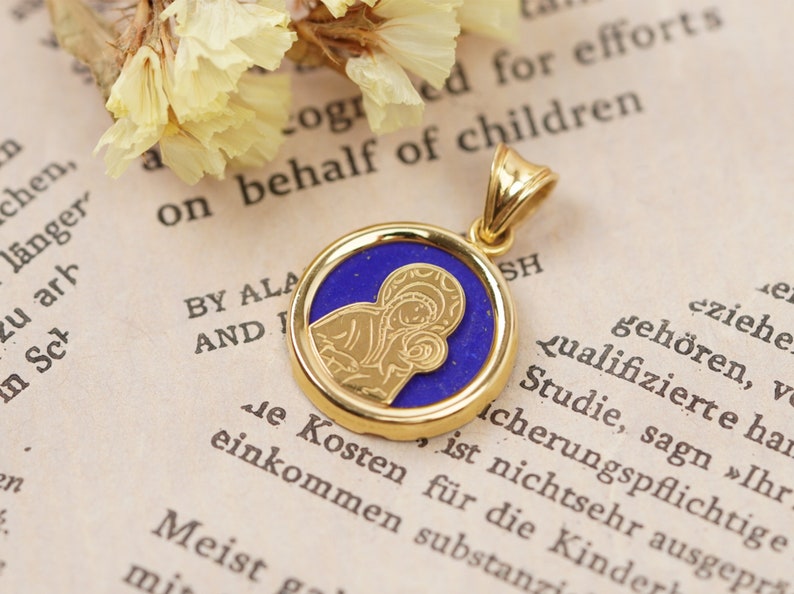 May include: Gold-tone pendant with a blue center featuring a religious icon. The pendant has a circular design with a detailed image of a religious figure. The pendant is on a vintage book page.