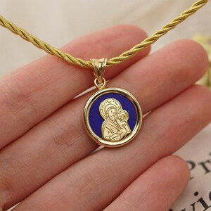 May include: Gold-tone pendant necklace featuring a round medallion with a blue center and a religious icon. The pendant hangs from a gold-colored cord. The necklace is a religious item.