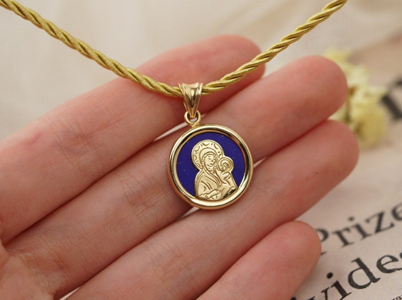 May include: Gold-tone pendant necklace featuring a round medallion with a blue center and a religious icon. The pendant hangs from a gold-colored cord. The necklace is a religious item.