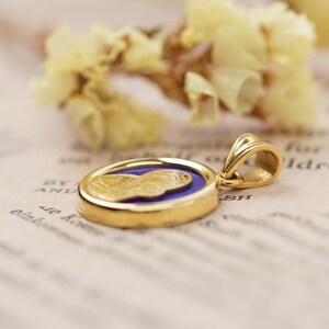 May include: Gold pendant with a detailed relief of a figure on a blue background. The pendant has a gold bail and is set against a soft, blurred background with dried flowers.
