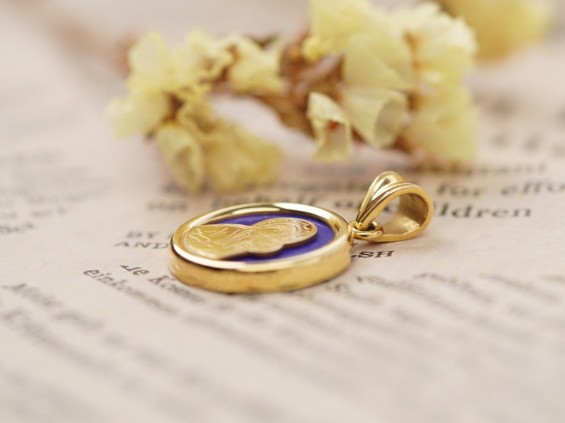 May include: Gold pendant with a detailed relief of a figure on a blue background. The pendant has a gold bail and is set against a soft, blurred background with dried flowers.